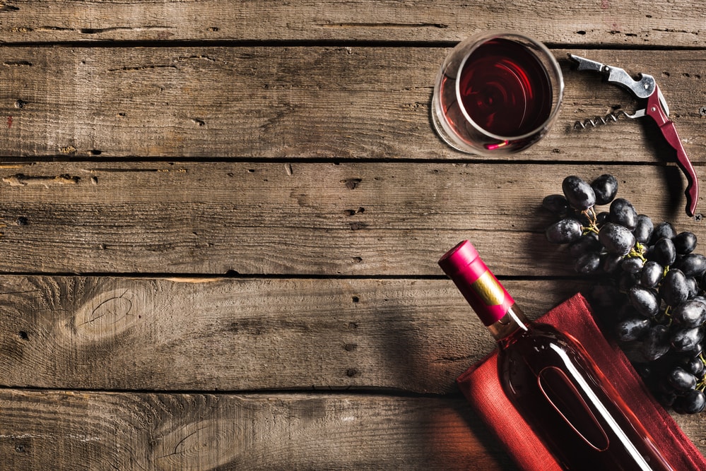 A bottle and glass of red wine laying on a wooden table, with a stem of grapes and a bottle open to the side