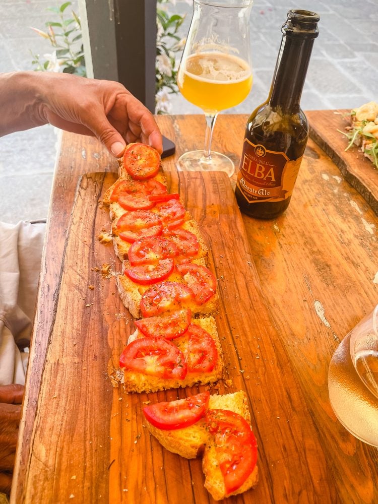 a long half loaf of bread covered with olive oil, tomatoes, and herbs served with an italian beer