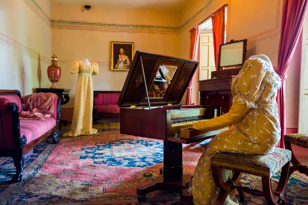 living room decorated in wooden tables including gold painted details, yellow carpet with a female dress sitting by piano and another standing across the room in the bolivar museum in bogota colombia