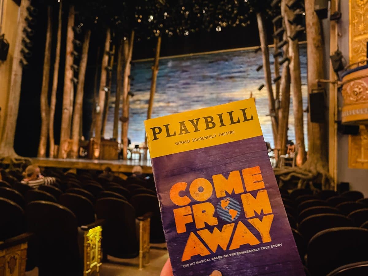 a playbill from the Broadway musical Come From Away with the theater and stage in the background