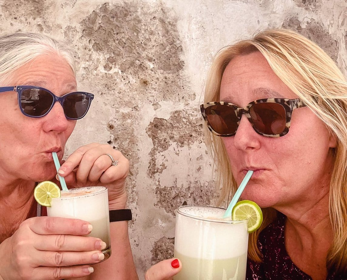 two women sipping on pisco sour cocktails against a terra cotta wall