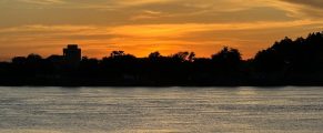Serene sunset over a river with silhouettes of trees and buildings in the distance.