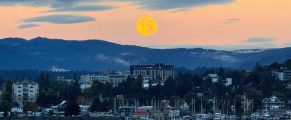 Moon over a scenic marina with boats and cityscape during sunset, travel destination in the Pacific Northwest.
