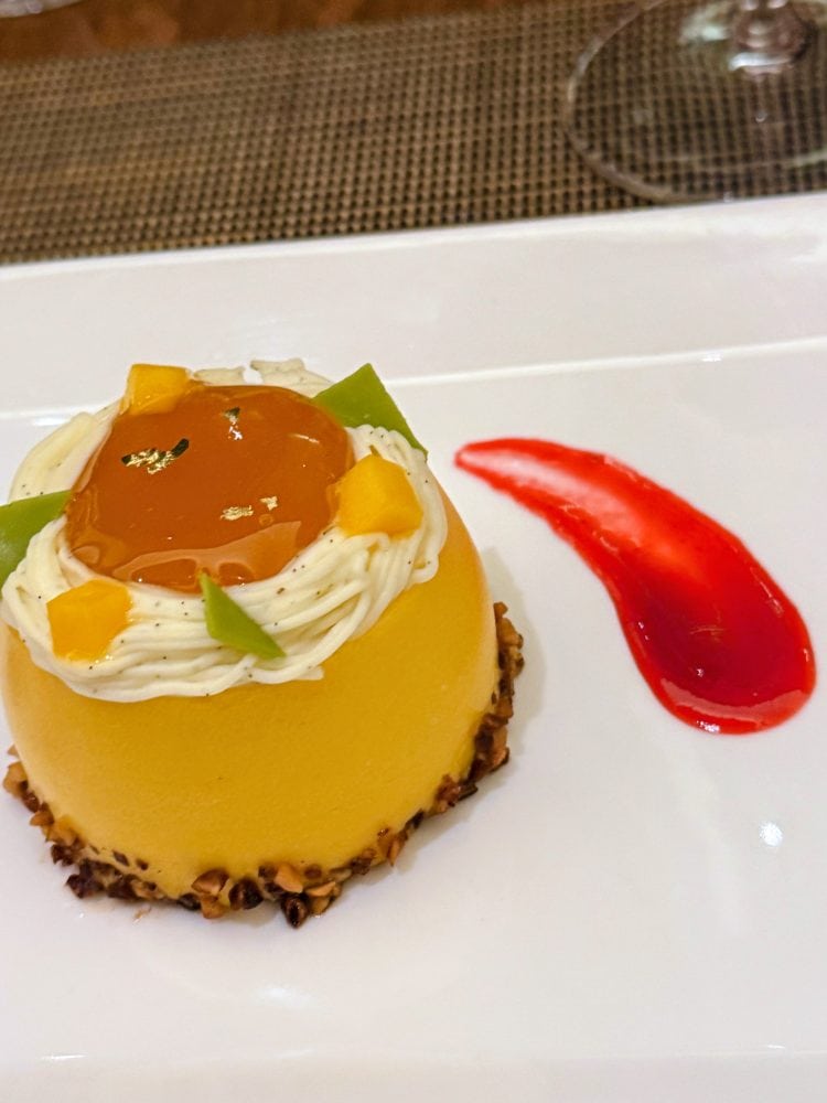 Elegant mango mousse dessert with fruit topping and red sauce on white plate.