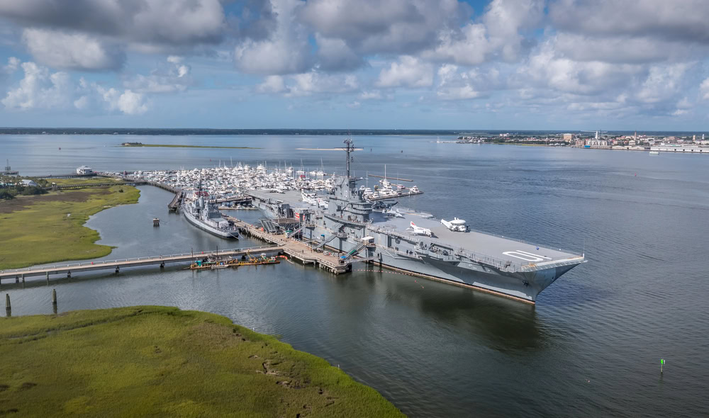 12 Terrific Museums in Charleston to Put on Your To-Visit List 5 Large naval ship docked at harbor with boats nearby and green marshland.